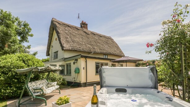 Ein Whirlpool mit einer Weinflasche und einem Glas, eine Terrasse mit einer Schaukel und im Hintergrund ein Haus mit einem Reetdach.