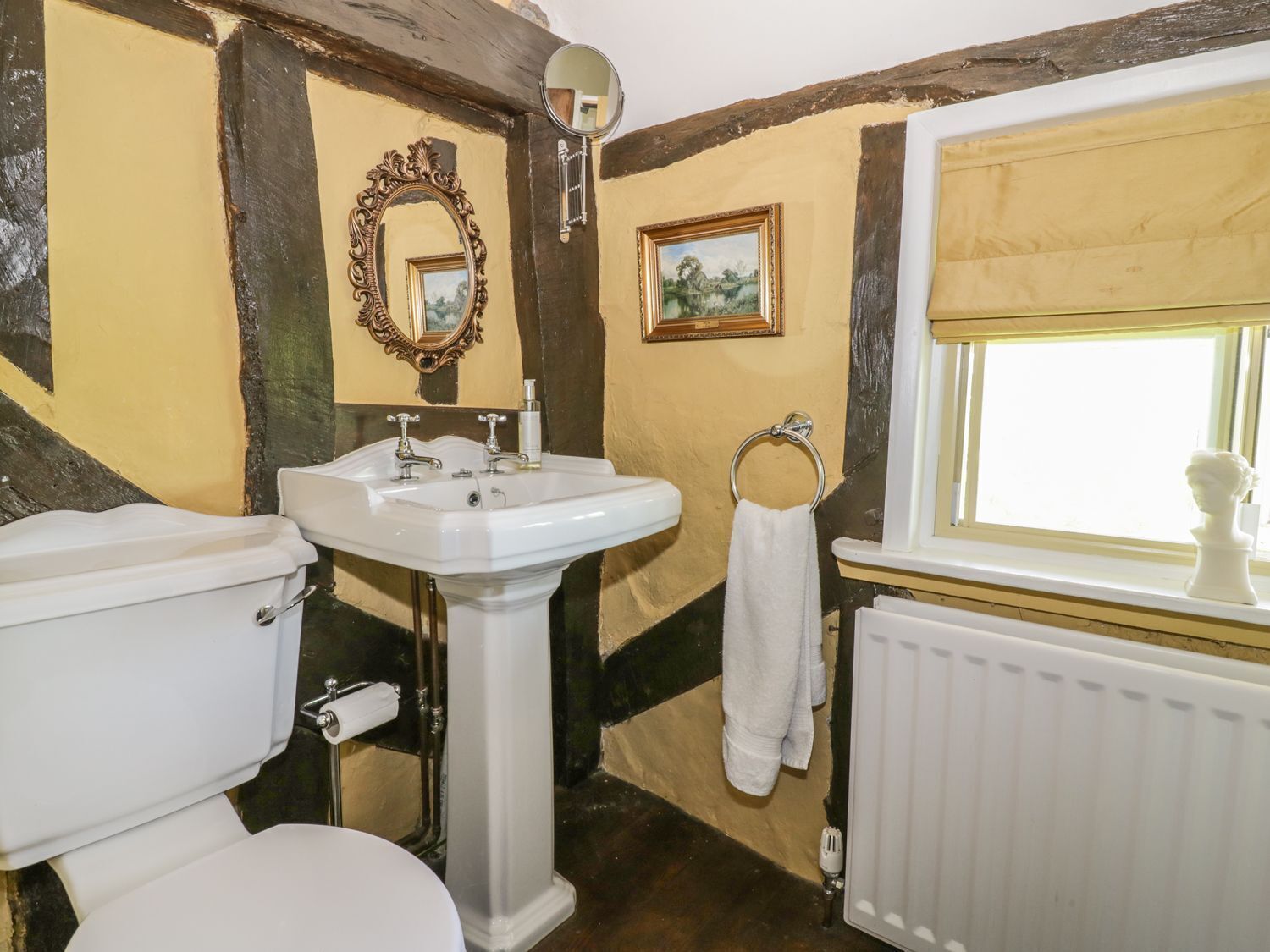 A bathroom with a white toilet, a pedestal sink, a mirror with an ornate frame, a towel rack, and a window with a view of the outside.