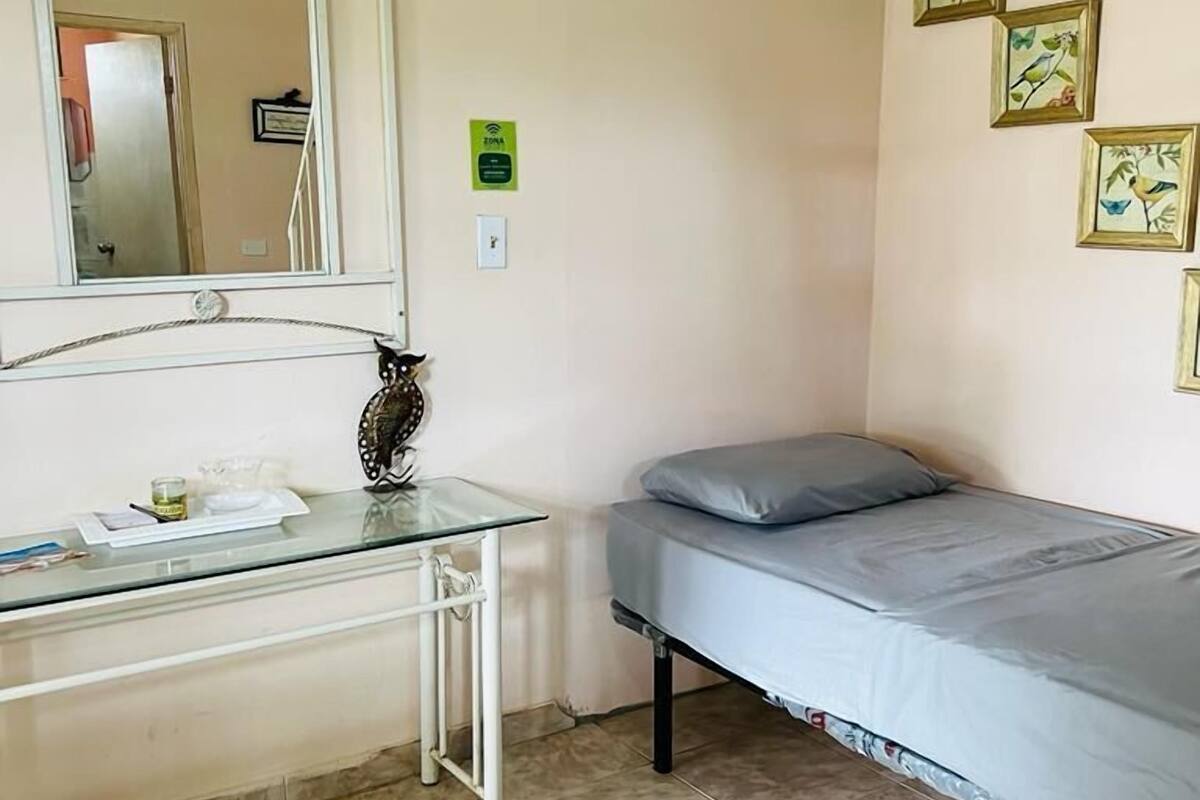 A small, neatly arranged room with a bed, a bedside table, a mirror, and a wall-mounted shelf.
