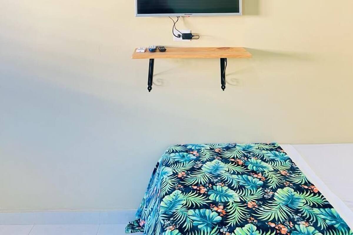 A room with a mounted television on a wall, a wooden table with a power strip, and a bed with a floral bedspread.
