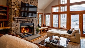 A cozy living room with a stone fireplace, a television, and a large window offering a mountain view. - Huge 7BR Mountain Lodge | Hot Tub, Fire Pit & Putting Green (Lincoln)