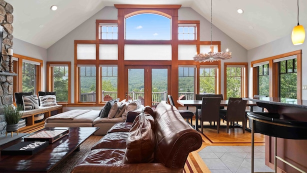 A spacious loft-style room with a high ceiling, large windows, a stone fireplace, a leather sofa, and a dining area. - Huge 7BR Mountain Lodge | Hot Tub, Fire Pit & Putting Green (Lincoln)