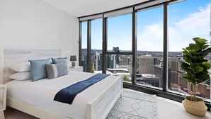 Un dormitorio moderno con una cama grande, una mesita de noche, una lámpara y vistas a la ciudad a través de amplias ventanas.