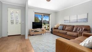 A modern living room with a brown leather sofa, a wooden coffee table with a flat-screen TV, a patterned rug, and a door with frosted glass panels. - Serenity Now Inverloch (Inverloch)