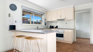 A modern kitchen with a central island, bar stools, a built-in oven, and a window overlooking the outside. - Serenity Now Inverloch (Inverloch)