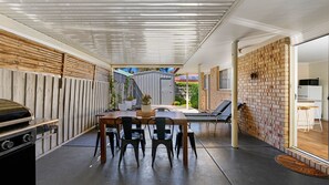 A covered outdoor dining area with a table and chairs, a grill, and a brick wall. - Serenity Now Inverloch (Inverloch)