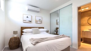 A modern bedroom with a large bed, a bedside lamp, a woven stool, and a mirror. - Elegant 2-Bed Apartment with Stunning Views (Batemans Bay)