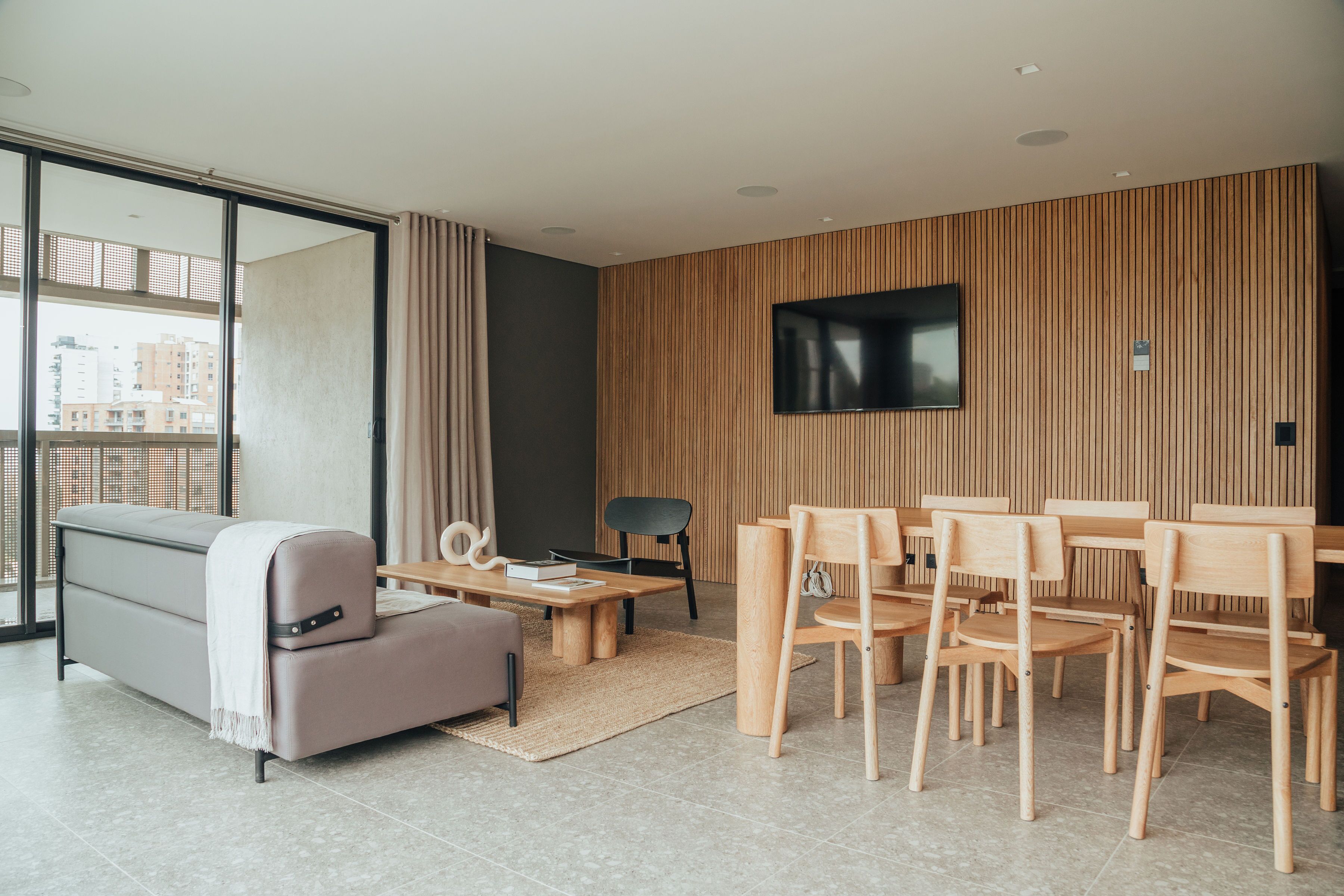 A modern living room with a sofa, coffee table, and dining area with wooden chairs and a TV mounted on the wall.