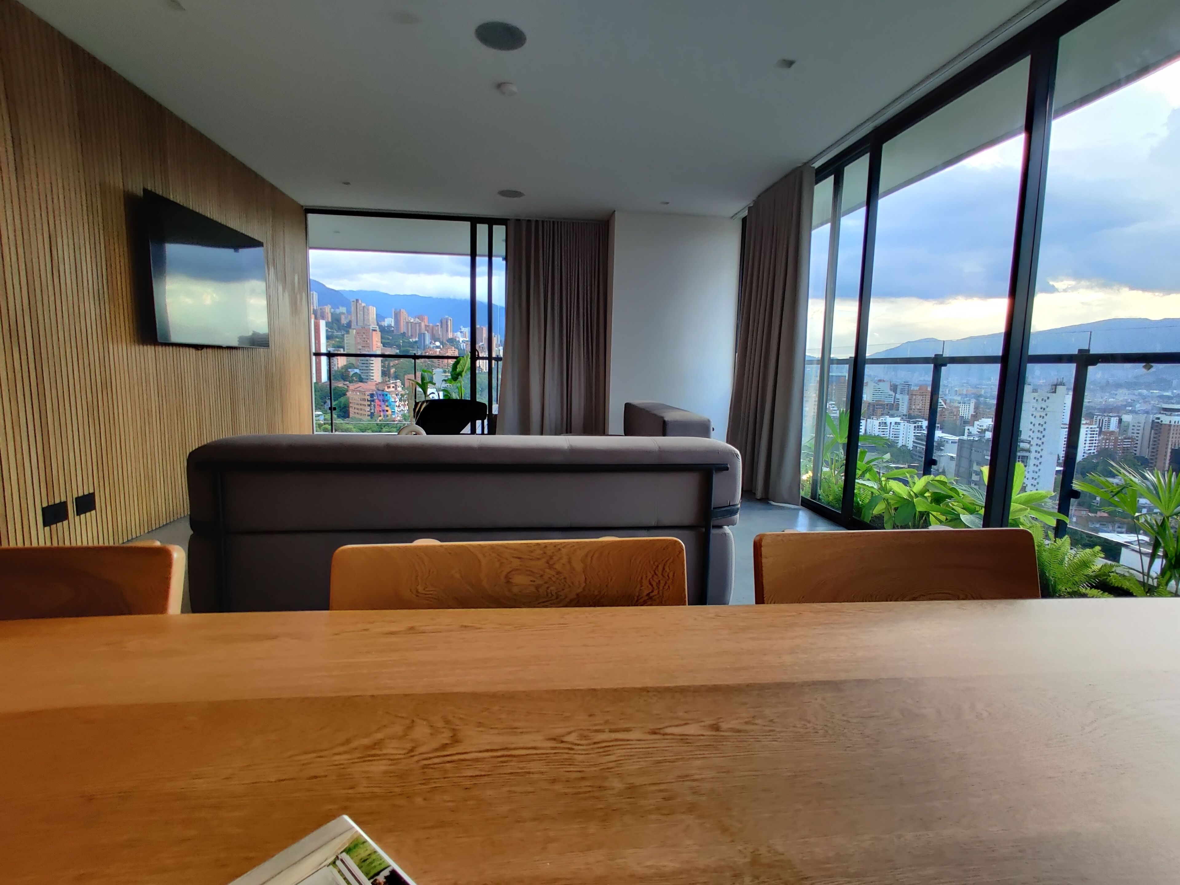 A modern room with a large window, a flat-screen TV mounted on the wall, and a dining area with a wooden table and chairs.