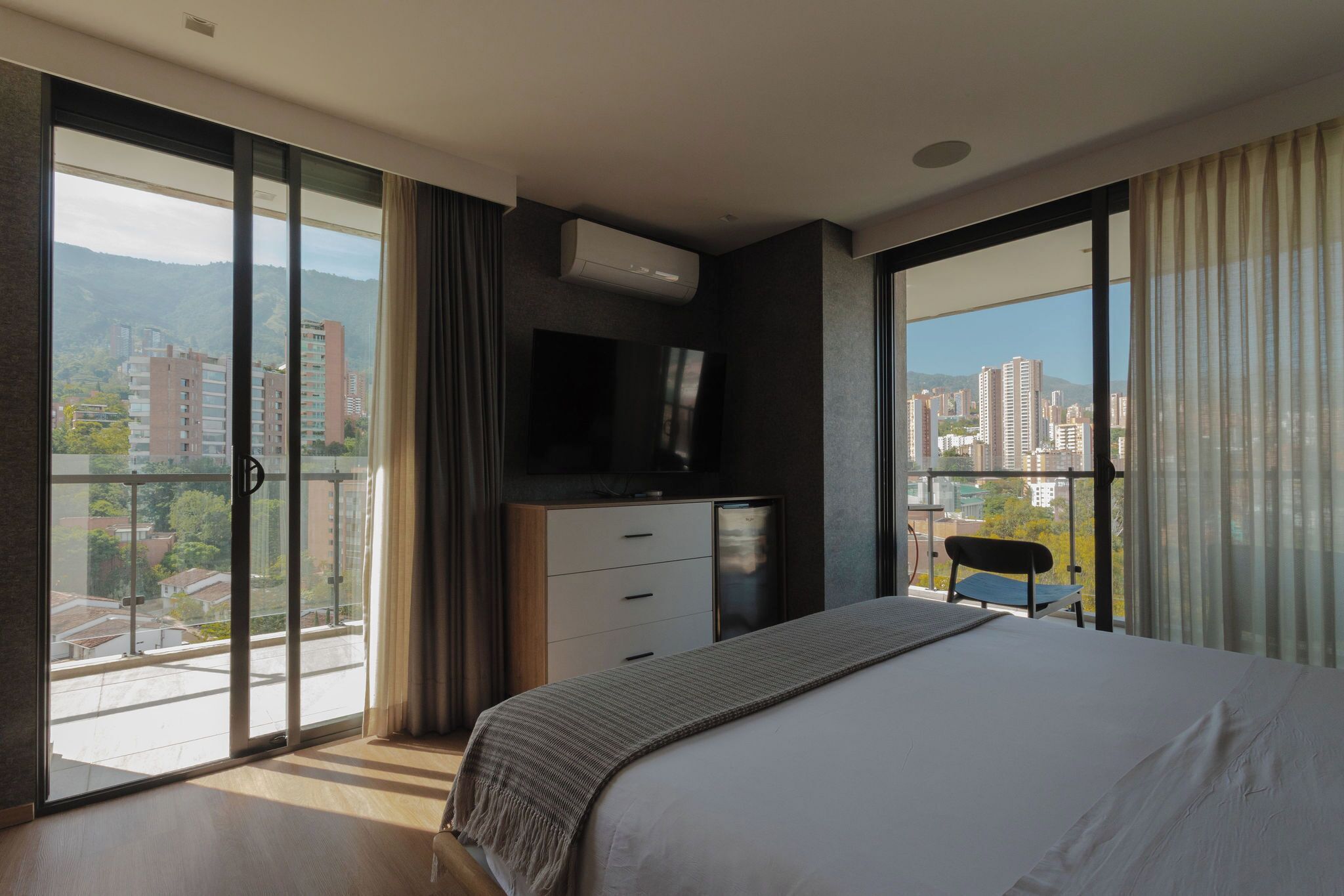 A modern bedroom with a large bed, a flat-screen TV, a dresser, and a balcony with a view of the city.