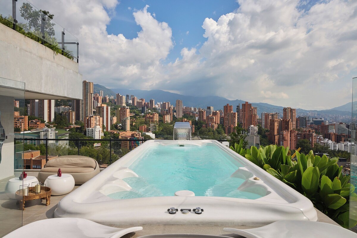 A rooftop infinity pool with a cityscape view, lounge seating, and a hot tub.