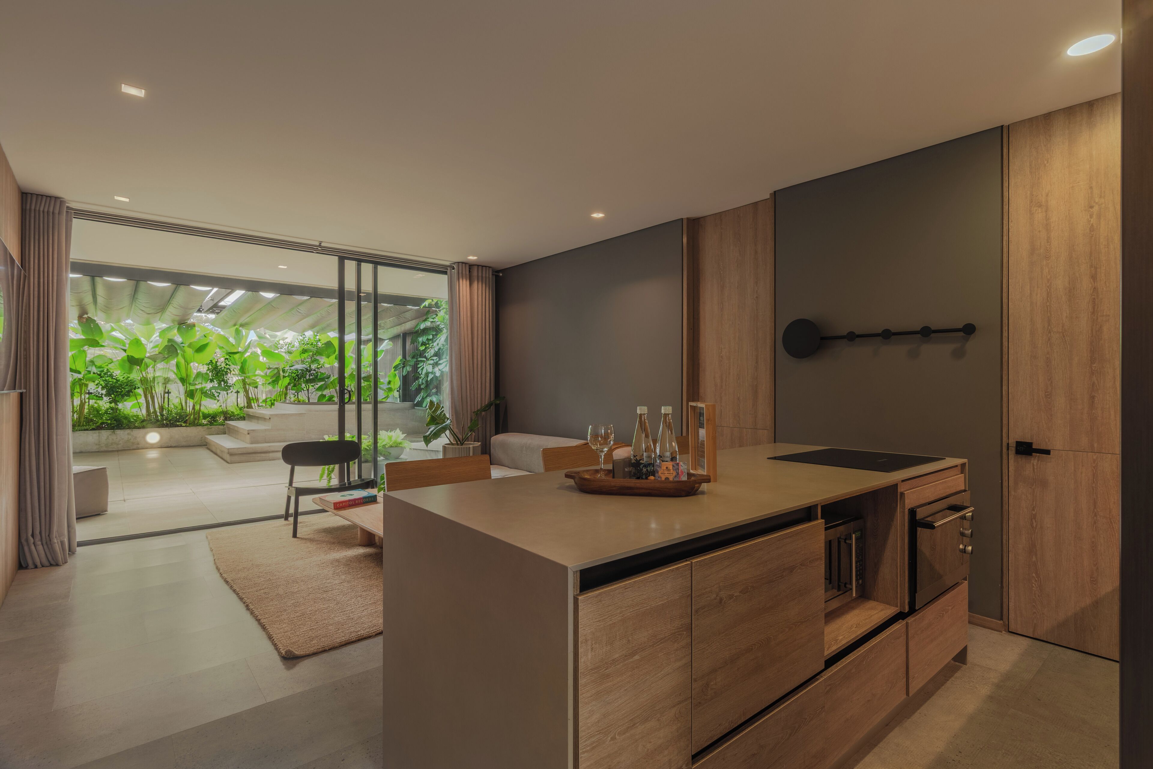 A modern kitchen with a central island, wooden cabinets, and a view of a green garden through large windows.