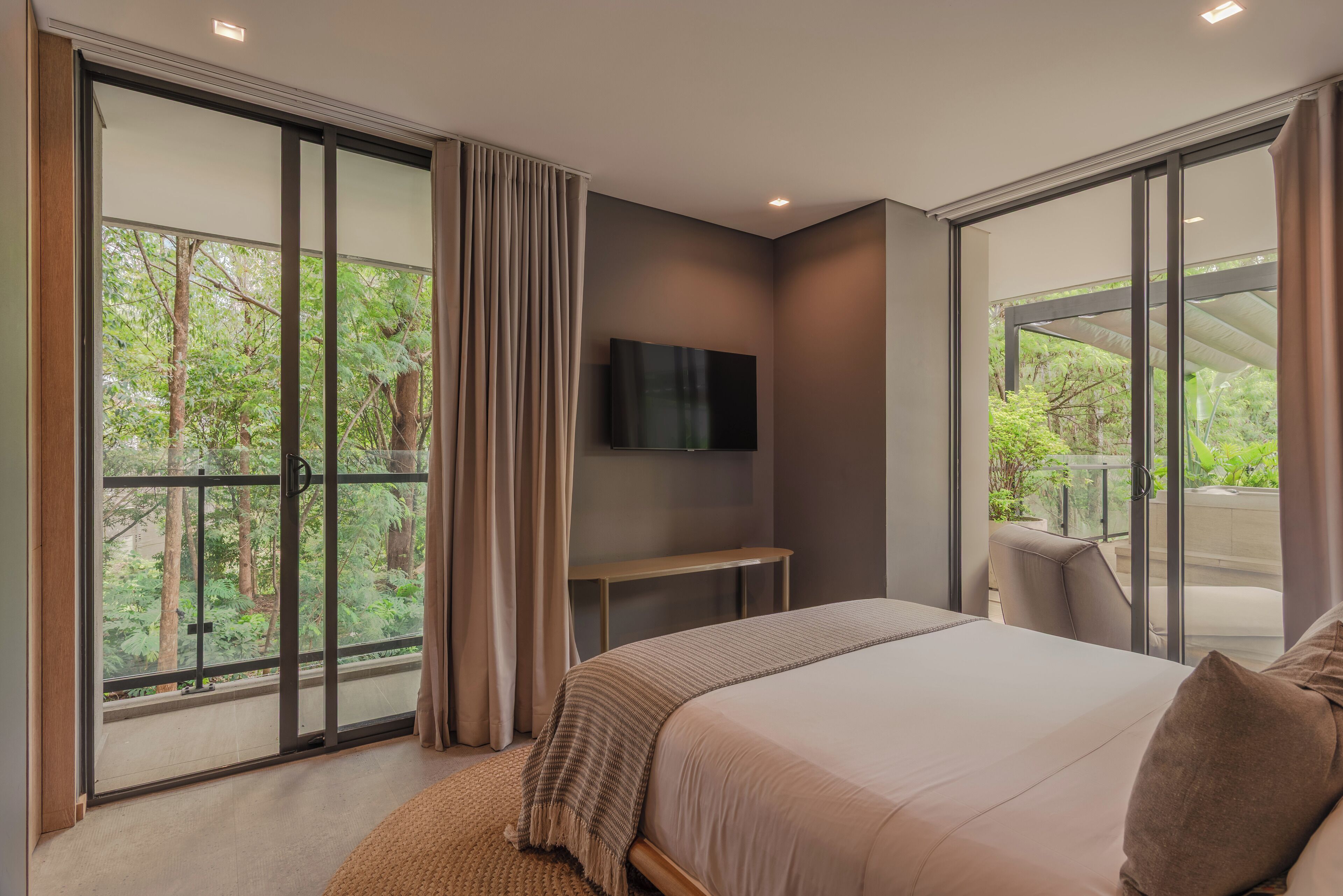A bedroom with a large sliding glass door to a balcony, a bed with a blanket, a TV mounted on the wall, and a small table.