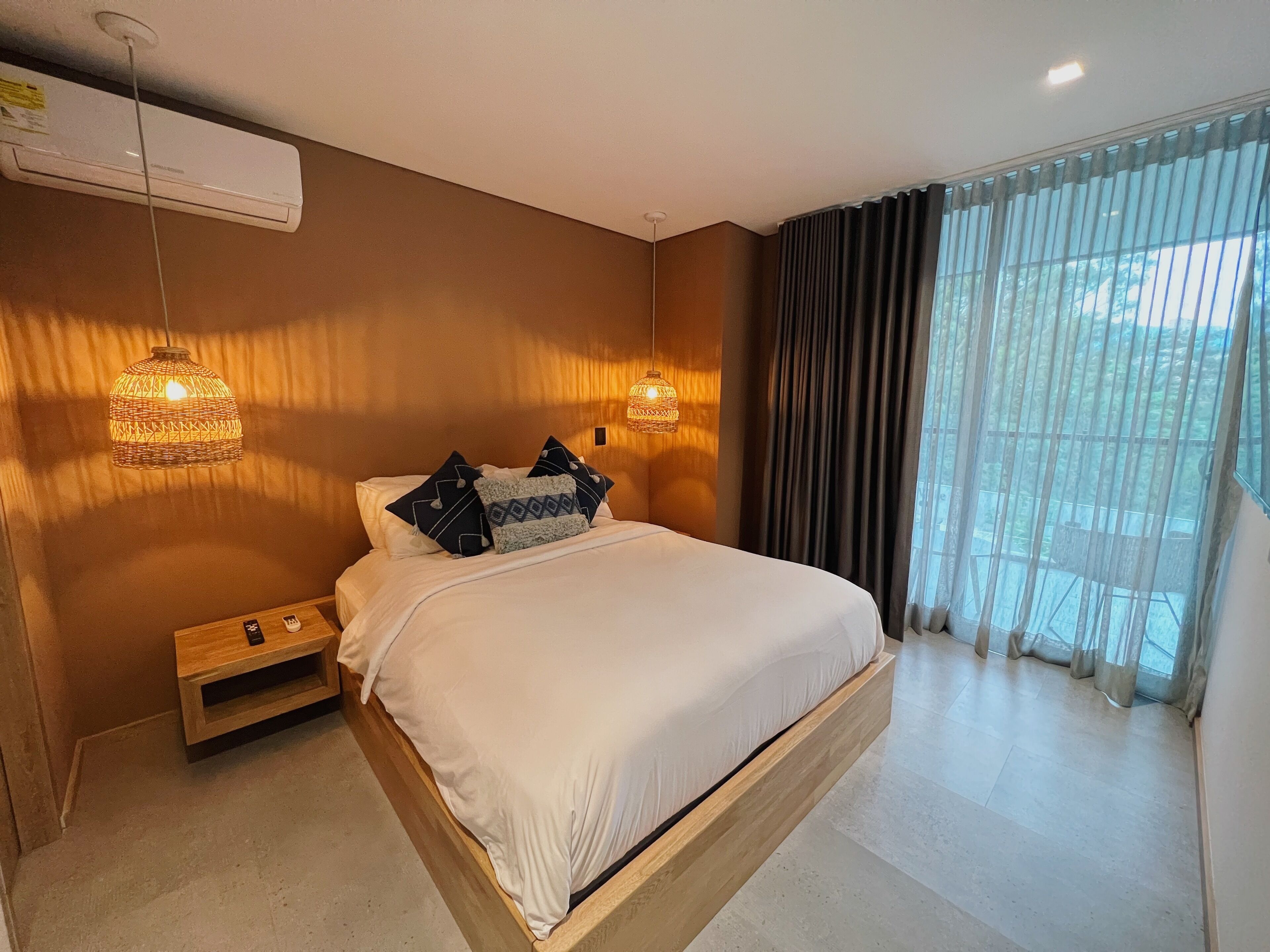 Une chambre moderne avec un grand lit, deux tables de chevet et un banc intégré. On y trouve deux lampes suspendues et une grande fenêtre avec des rideaux.