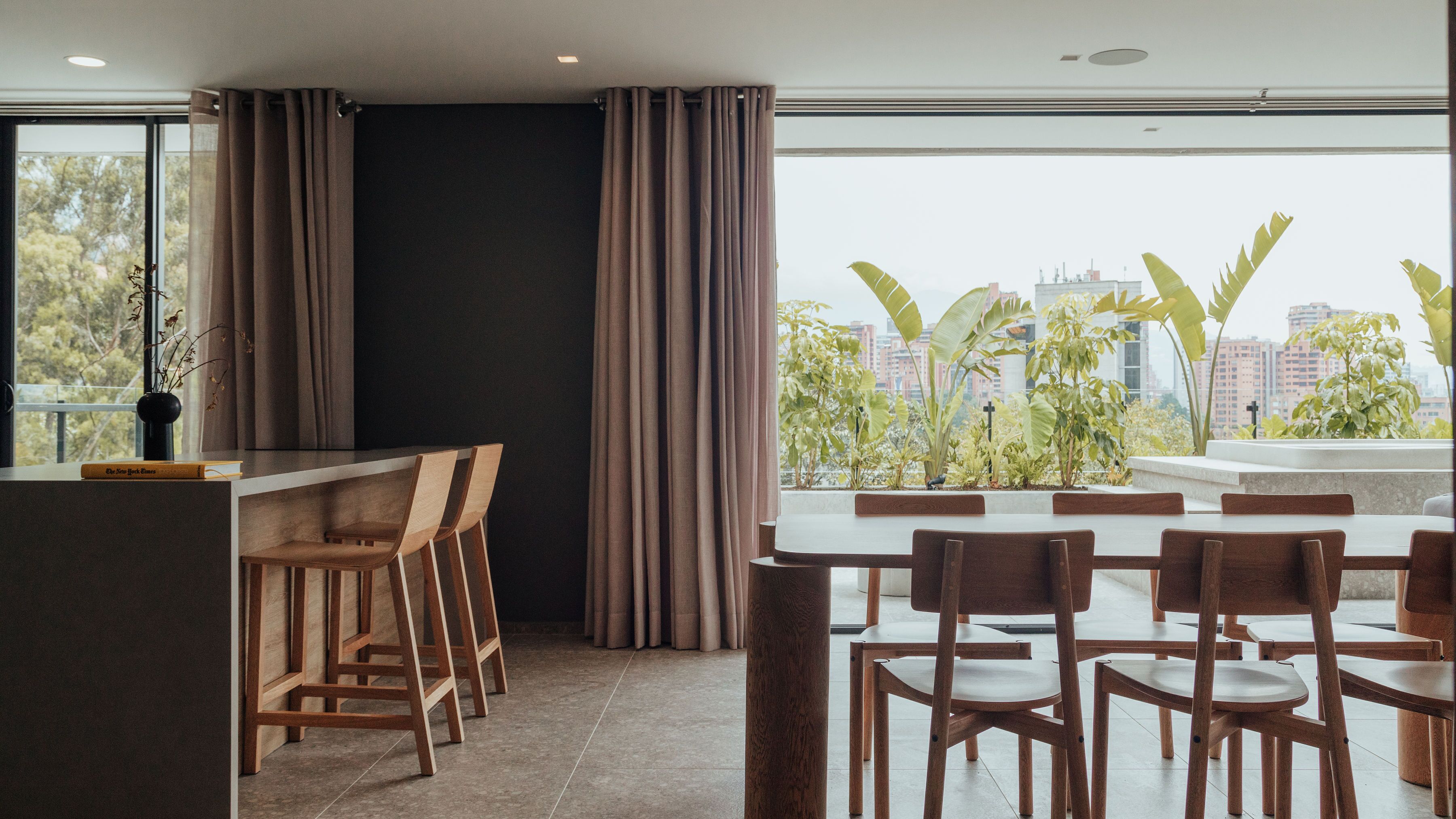 A modern dining area with a high ceiling, large windows, and a view of a cityscape.