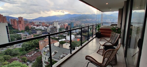 Un balcón con mobiliario de mimbre y vista a un paisaje urbano con montañas al fondo.