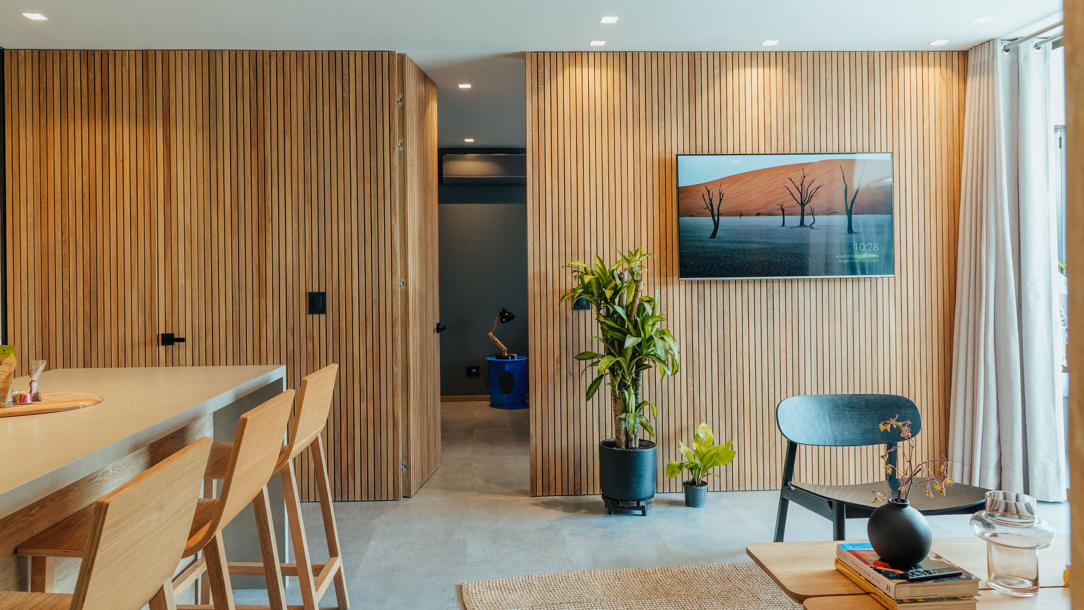 Un interior moderno con un panel de madera, una zona de comedor con sillas de madera, una mesita auxiliar azul, una planta en maceta, una silla negra y un cuadro en la pared.