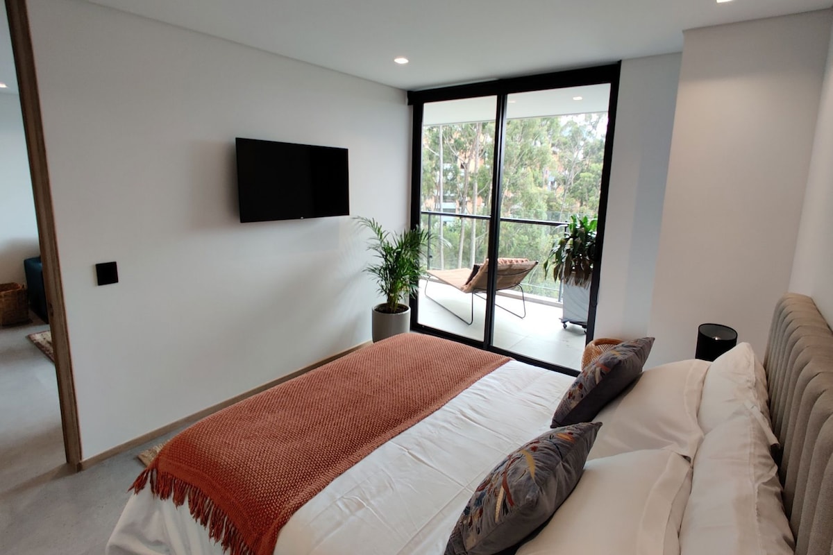 A modern bedroom with a large bed, a flat-screen TV mounted on the wall, and a balcony with a view of trees.