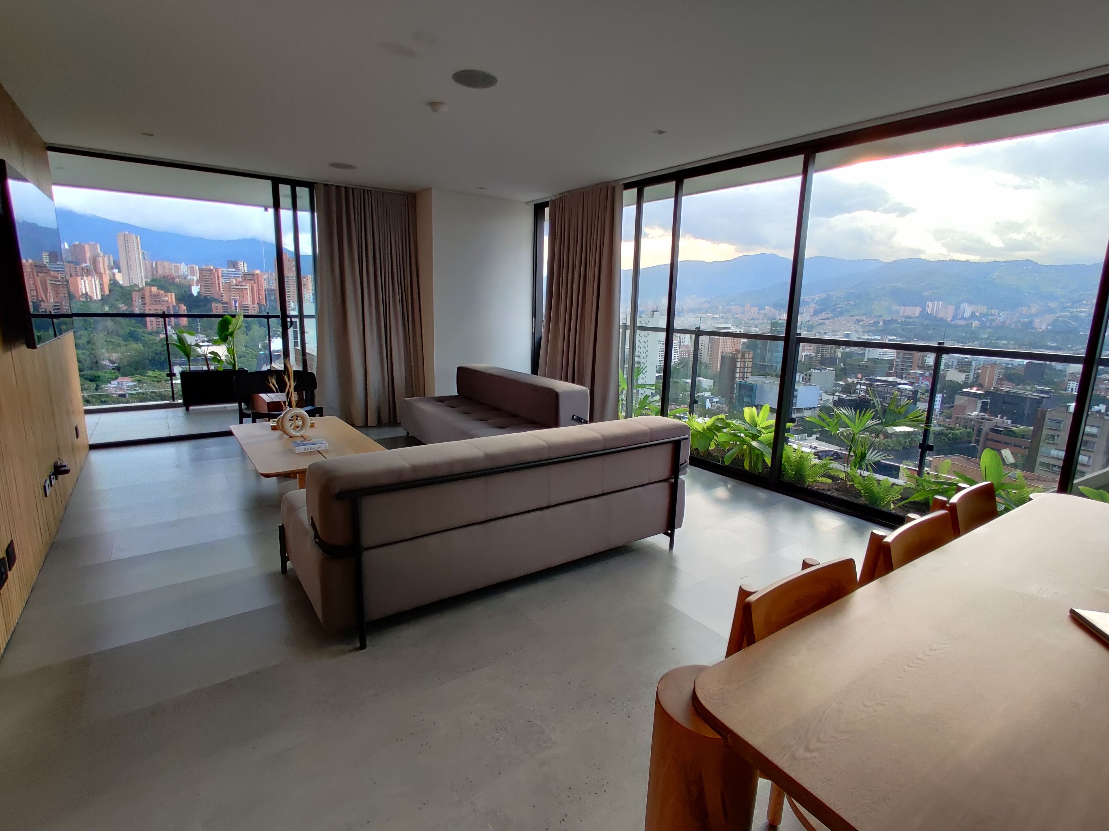 A spacious living area with a sofa, dining table, and chairs, large windows offering a city view.