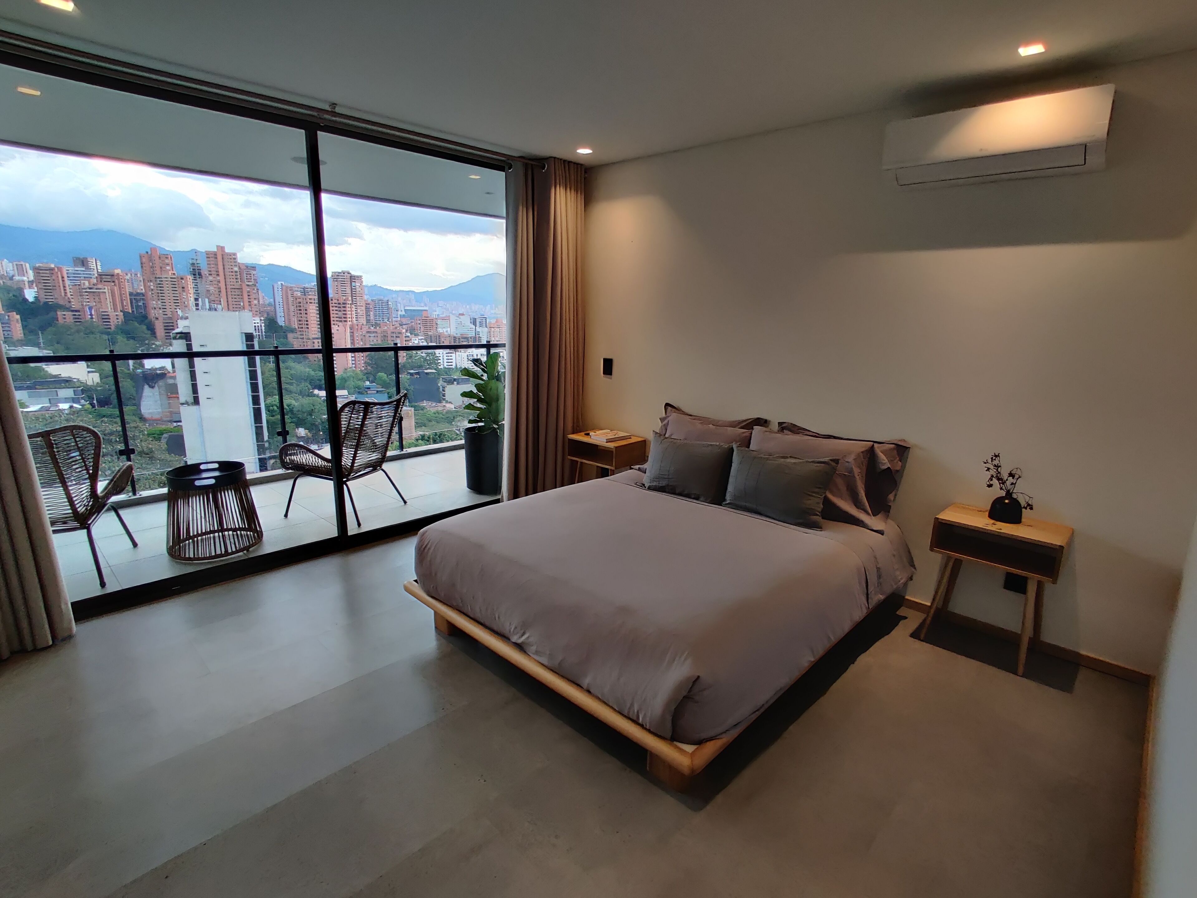 A modern bedroom with a large bed, a nightstand, a potted plant, and a view of a cityscape through floor-to-ceiling windows.
