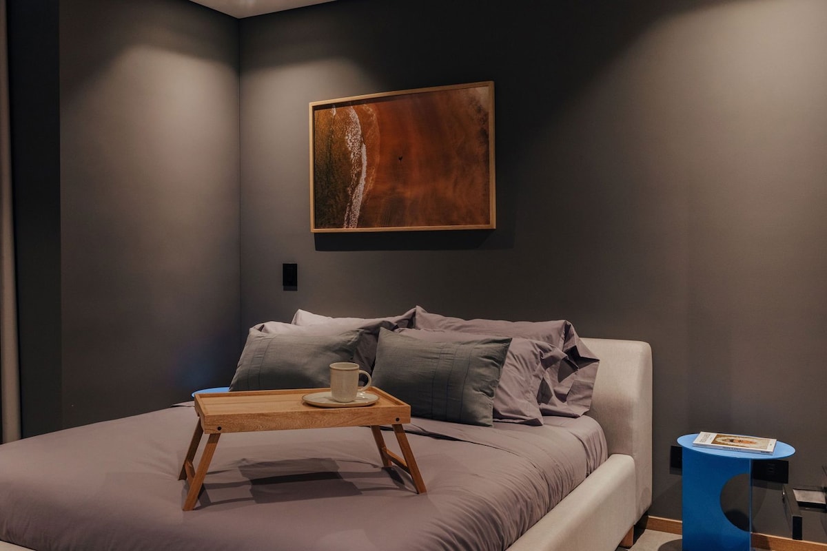 A modern bedroom with a bed, a bedside table with a cup, a painting on the wall, and a blue side table.