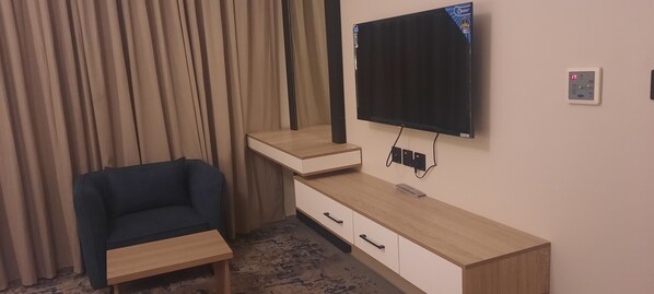 A room with a TV mounted on the wall, a wooden desk with drawers, a blue chair, and a curtain. - Golden New Quraish (Jeddah)