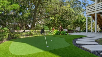 Een goed onderhouden midgetgolfbaan met een putting green, omgeven door weelderige begroeiing en een terras met buitenmeubilair.
