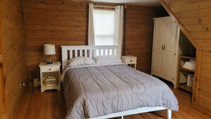 A bedroom with a wooden floor, a bed with a white headboard, a nightstand with a lamp, a window with blinds, and a wardrobe. - Sleeps 10, close to shopping centers on quiet dead end street. (Brunswick)