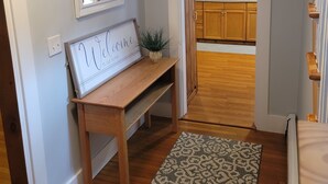A hallway with a wooden bench, a welcome sign, a small plant, and a patterned rug leading to a kitchen with wooden cabinets and a sink. - Sleeps 10, close to shopping centers on quiet dead end street. (Brunswick)