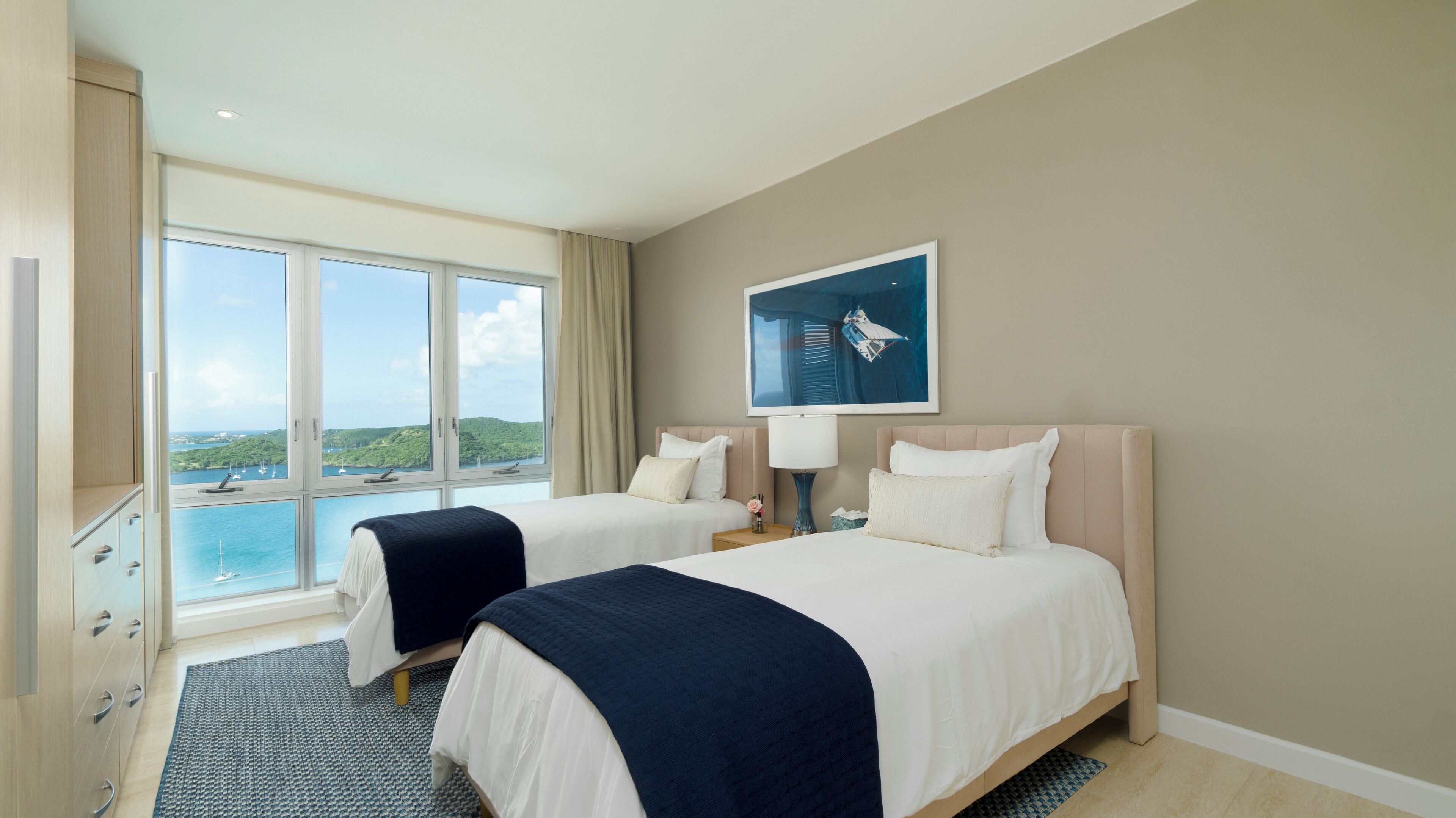 A hotel room with two beds, a large window with a view of the sea, and a painting on the wall.