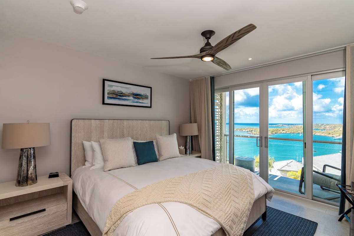 A hotel room with a large bed, two bedside tables, a ceiling fan, and a view of the ocean.