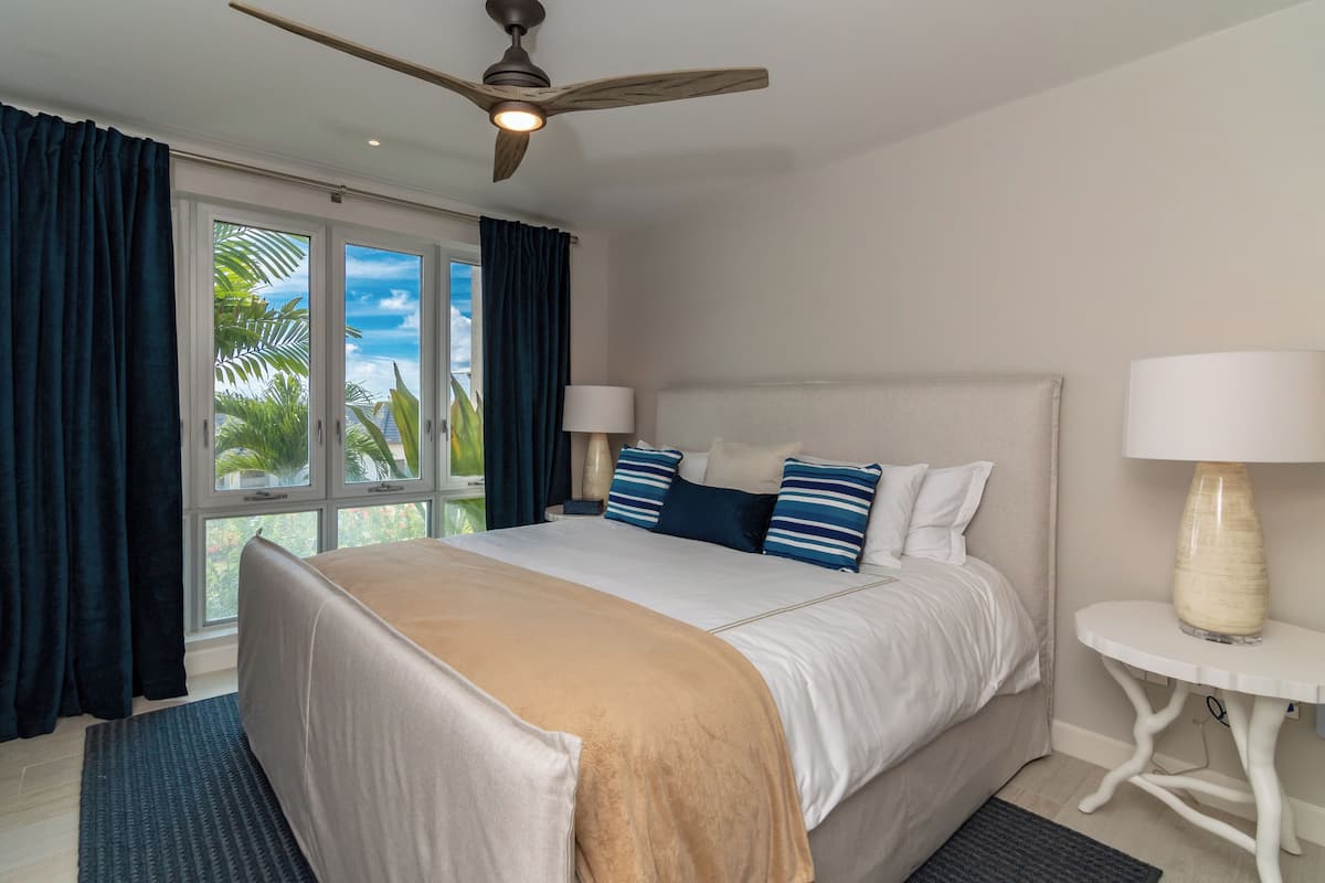 A hotel room with a large bed, a ceiling fan, a window with curtains, and a small table with a lamp.