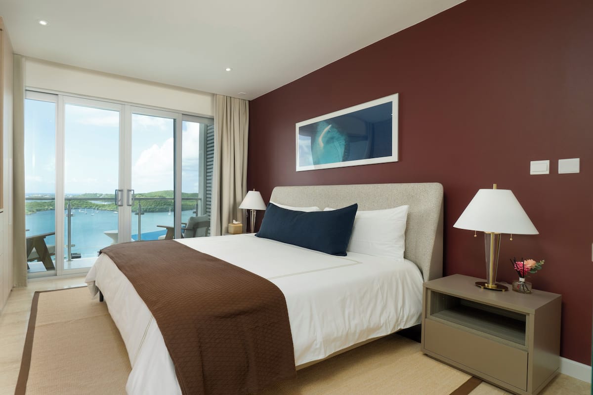 A hotel room with a large bed, two bedside lamps, a view of the sea, and a balcony with a table and chairs.