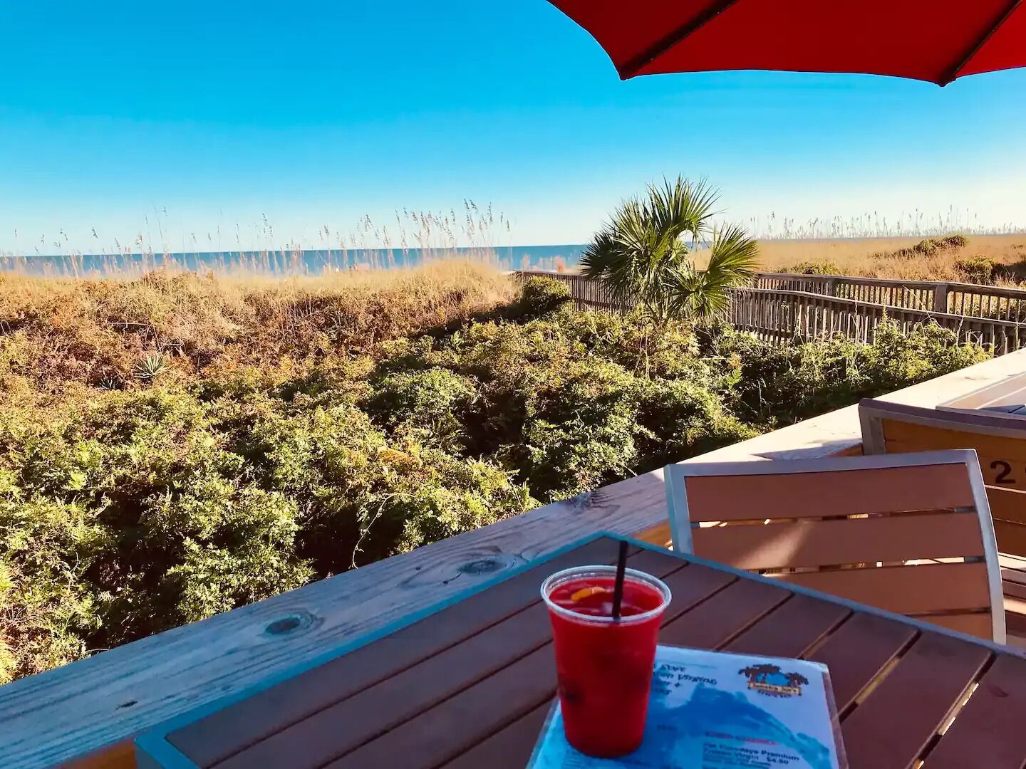 Mesa com bebida vermelha e cardápio, com vista para uma praia com grama seca e cerca de madeira.