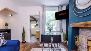 Uma sala de estar moderna com um sofá azul, uma mesa de jantar de vidro com duas cadeiras, uma lareira e uma janela grande com cortinas.