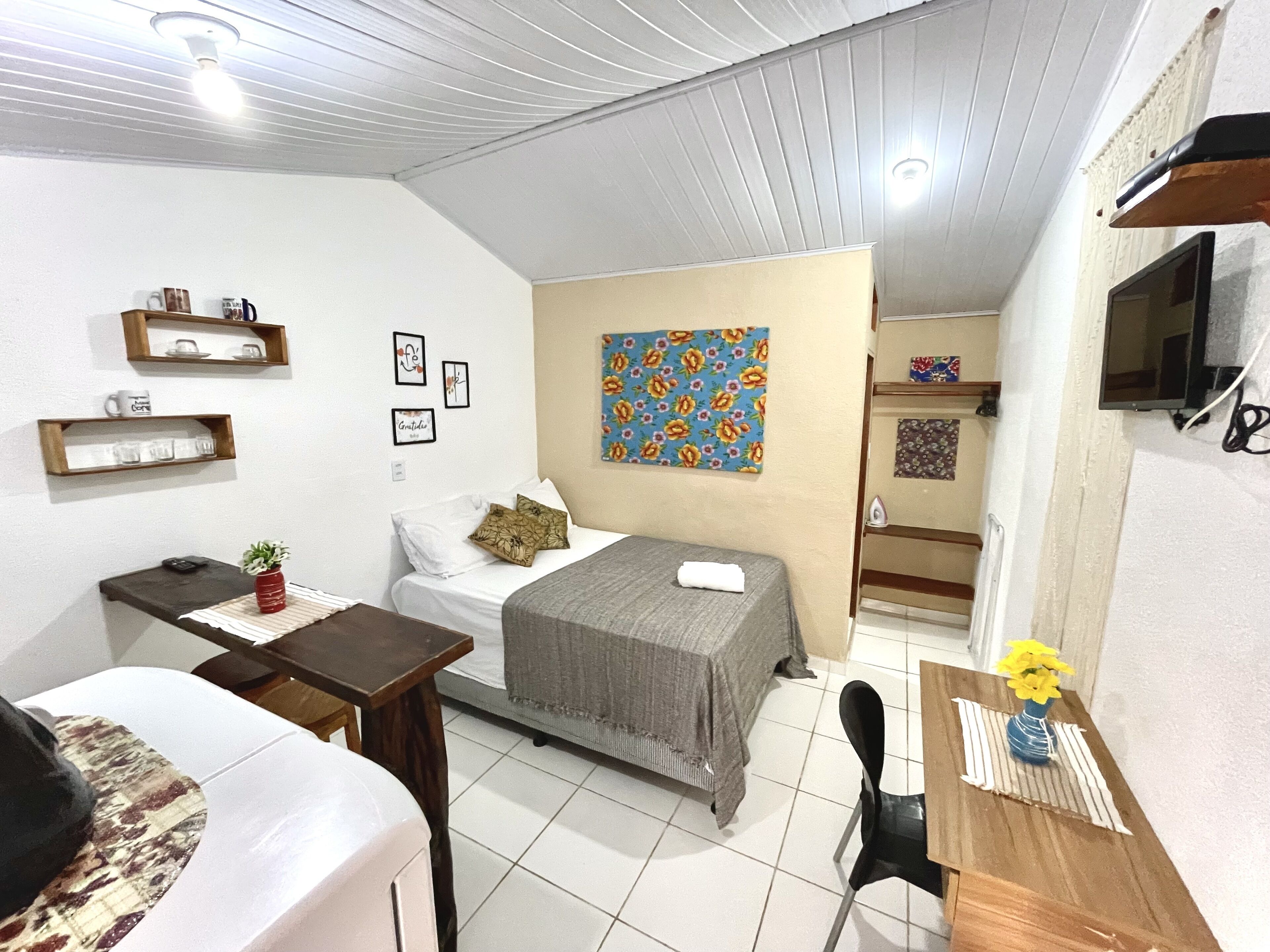 A compact hotel room with a bed, bedside table, a small desk, a chair, a TV, and a bathroom visible through an open door.