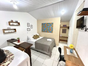 A compact hotel room with a bed, bedside table, a small desk, a chair, a TV, and a bathroom visible through an open door.