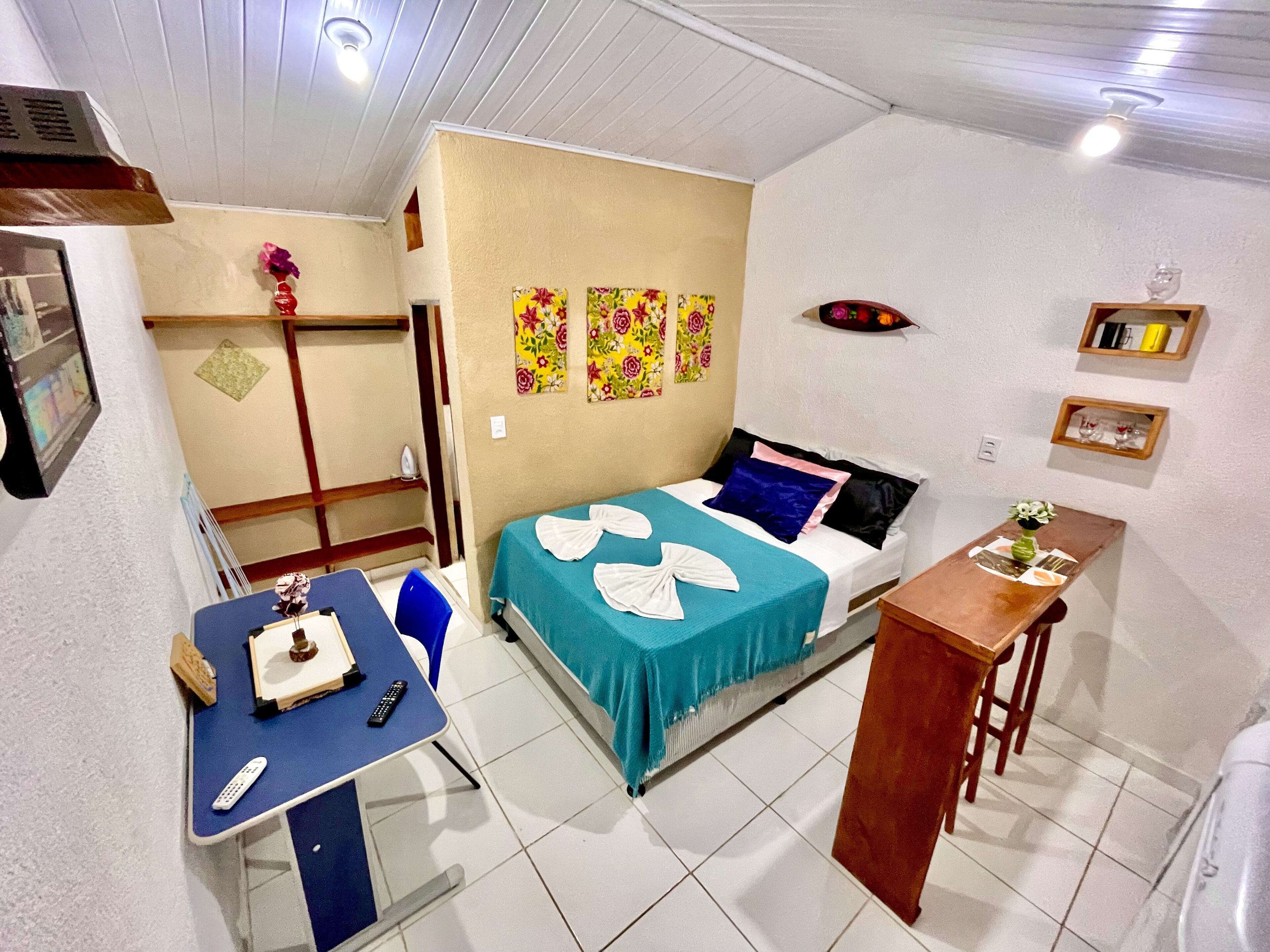 Habitación de hotel compacta con una cama, un escritorio con silla, una mesita pequeña y una estantería con adornos.