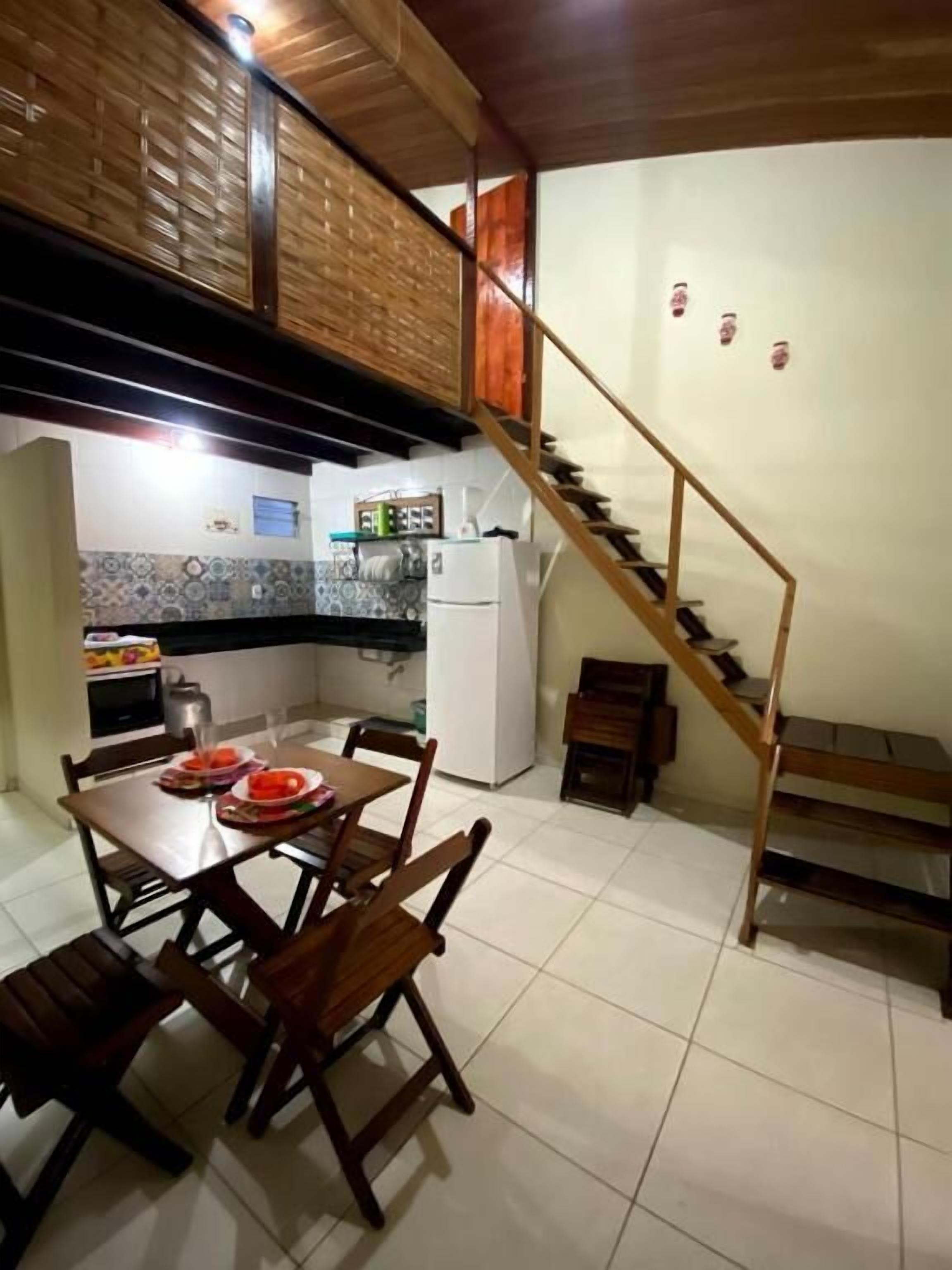 Un comedor con mesa y sillas de madera, una cocina con refrigerador y estufa, y una escalera de madera.