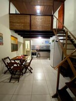 Uma sala de jantar com uma mesa e cadeiras de madeira, uma cozinha com frigorífico e fogão, e uma escadaria de madeira.