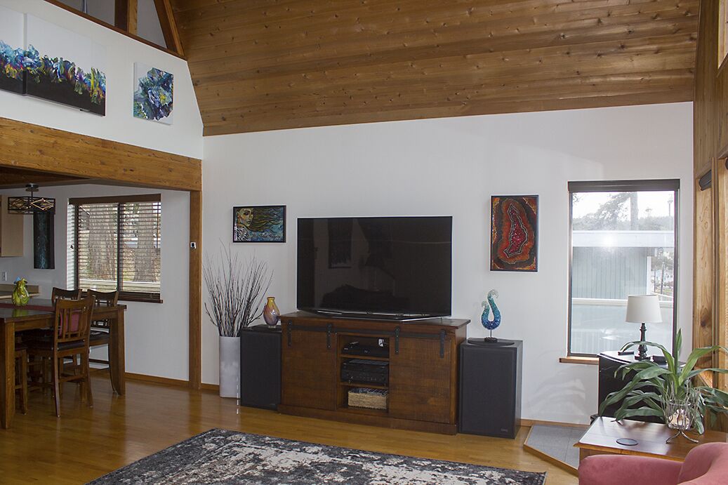 A spacious living room with a wooden ceiling, a large flat-screen TV on a wooden stand, a dining area with a table and chairs, and a large window providing natural light.