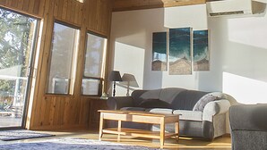 Un salon spacieux avec un plafond haut, des poutres apparentes, de grandes fenêtres, un ventilateur de plafond, un canapé, une table basse et des œuvres d’art accrochées au mur.
