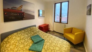Un dormitorio con una cama, un sillón amarillo, un reposapiés rojo, un ventanal grande y un televisor montado en la pared.