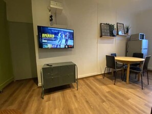 Una sala moderna con un televisor de pantalla plana montado en la pared, un mueble bajo, una mesa de comedor con sillas y un refrigerador.