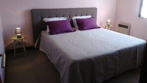 A bedroom with a large bed, two nightstands, a window, and a wall-mounted heater. - Gite with large swimming pool, play area, green space.  (Saint-Marcory)