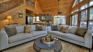 A cozy cabin interior with two grey sofas, a round coffee table, and a kitchen area with cabinets and a refrigerator. - Lideca’s Treehouse | Hot Tub + Fire Pit + Games (Mineral Bluff)