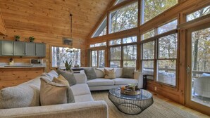 A cozy cabin interior with a large sofa, a round coffee table, and a kitchen area with cabinets and a microwave. - Lideca’s Treehouse | Hot Tub + Fire Pit + Games (Mineral Bluff)