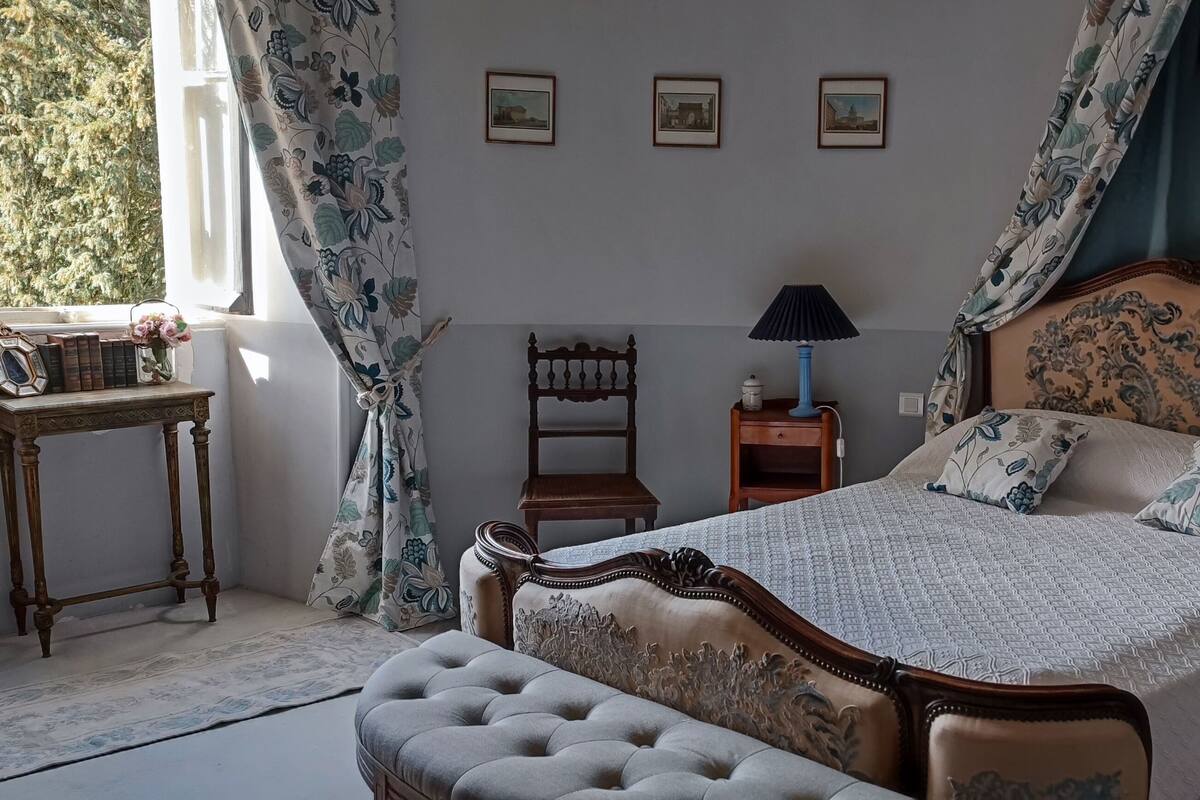 A bedroom with a large bed, a bedside table, a nightstand, and a window with curtains.