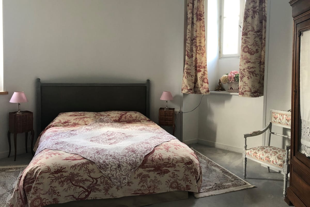 A bedroom with a bed, two bedside tables with lamps, a chair, a window with curtains, and a small shelf with flowers.