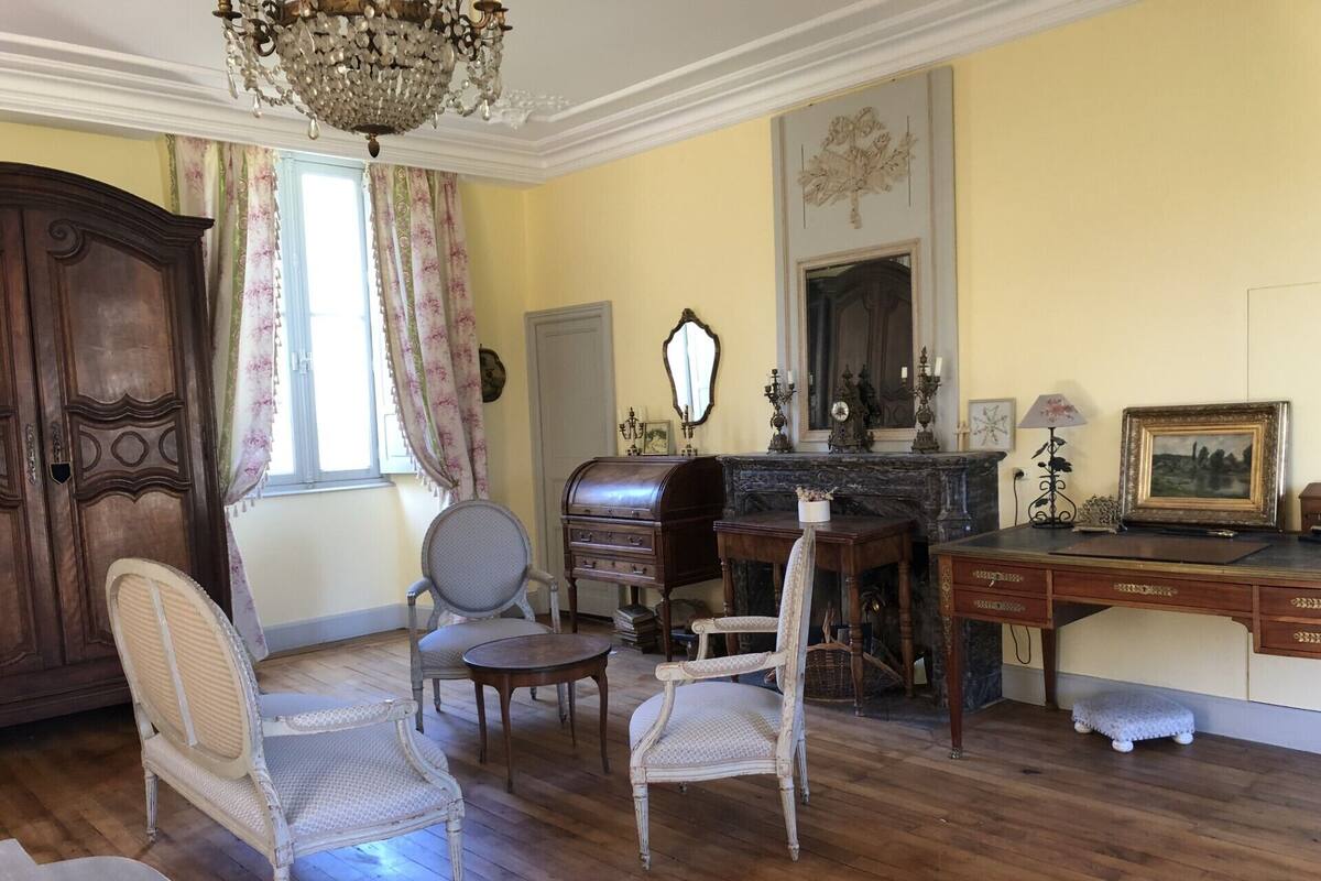 A room with a wooden floor, a chandelier, a large wooden cabinet, a desk, a chair, and a fireplace.
